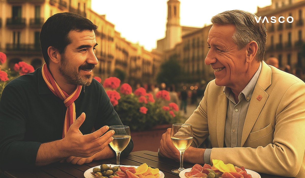 Двама мъже се наслаждават на напитки и тапас в открито кафене на испански площад, разговаряйки с цветя и архитектура на заден план.