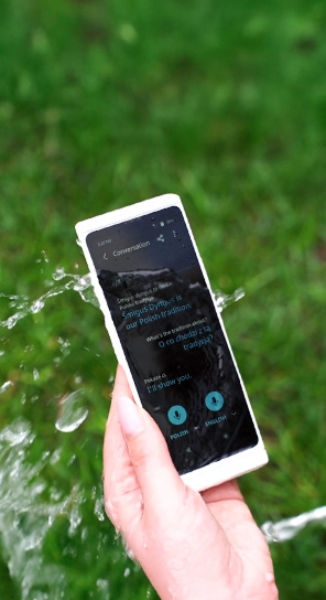 Близък план на ръка, държаща Vasco Translator V4, докато водата пръска устройството, подчертавайки неговата издръжливост.