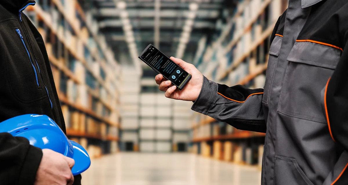 Двама души стоят един срещу друг; единият държи каска, а другият – Vasco Translator V4. На екрана на устройството се вижда активен интерфейс за превод на реч.