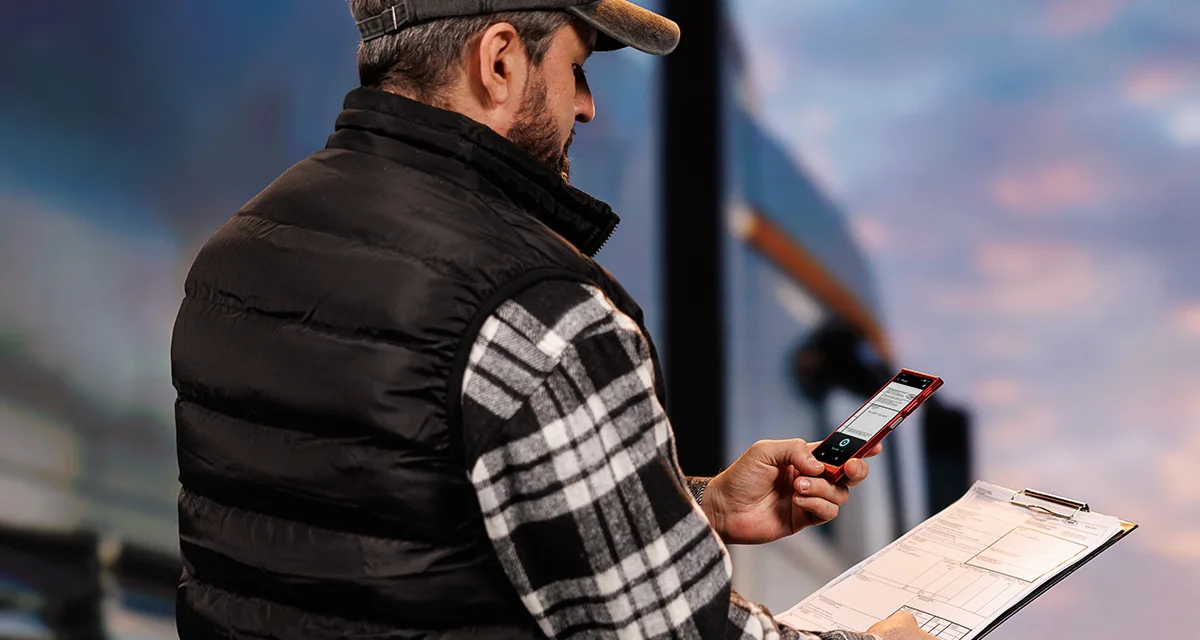 Шофьор на камион стои пред камиона, като в едната ръка държи документи, а в другата – Vasco Translator V4. На екрана на устройството е показана функцията за превод на снимки.