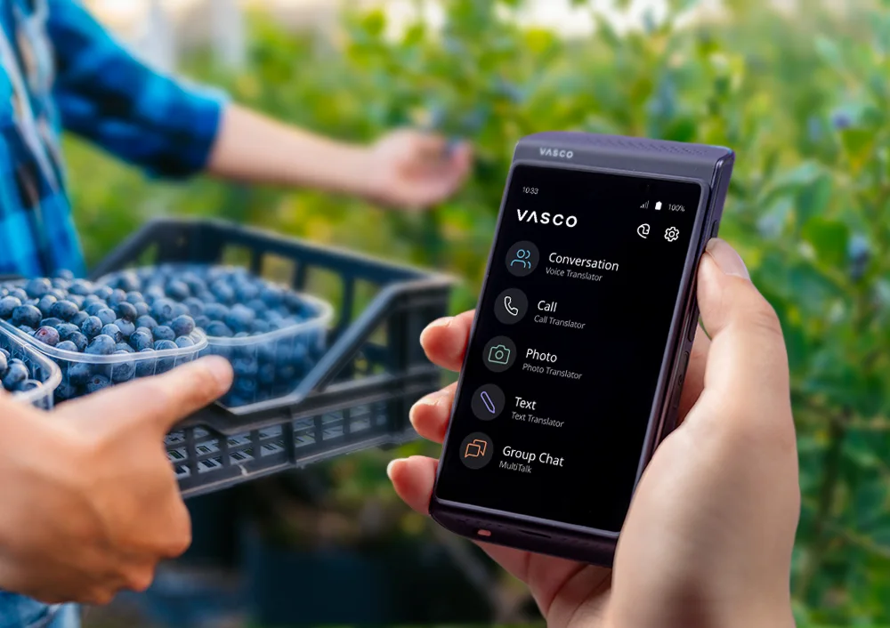 Vasco Translator Q1 на преден план, с боровинкови храсти на заден план и човек, държащ щайга с набрани боровинки.
