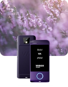 Изображение за представяне на продукта, показващо лилаво устройство „Vasco Translator“ от два ъгъла на фона на тъмна текстурирана тъкан. Устройството вляво е показано отзад, като се виждат обективът на камерата със светкавица и релефното лого „VA“. Устройството вдясно показва предната част с осветен екран, на който се виждат поздравите „Hello!“, „VA“, „¡Hola!“ и логото „VASCO Translator“. Кръгъл бутон в долната част на предната страна на устройството е очертан със сияйна синя светлина. Композицията е разположена на фона на мечтателен, замъглен фон от лилави цветя от вереск, облъчени от мека слънчева светлина.